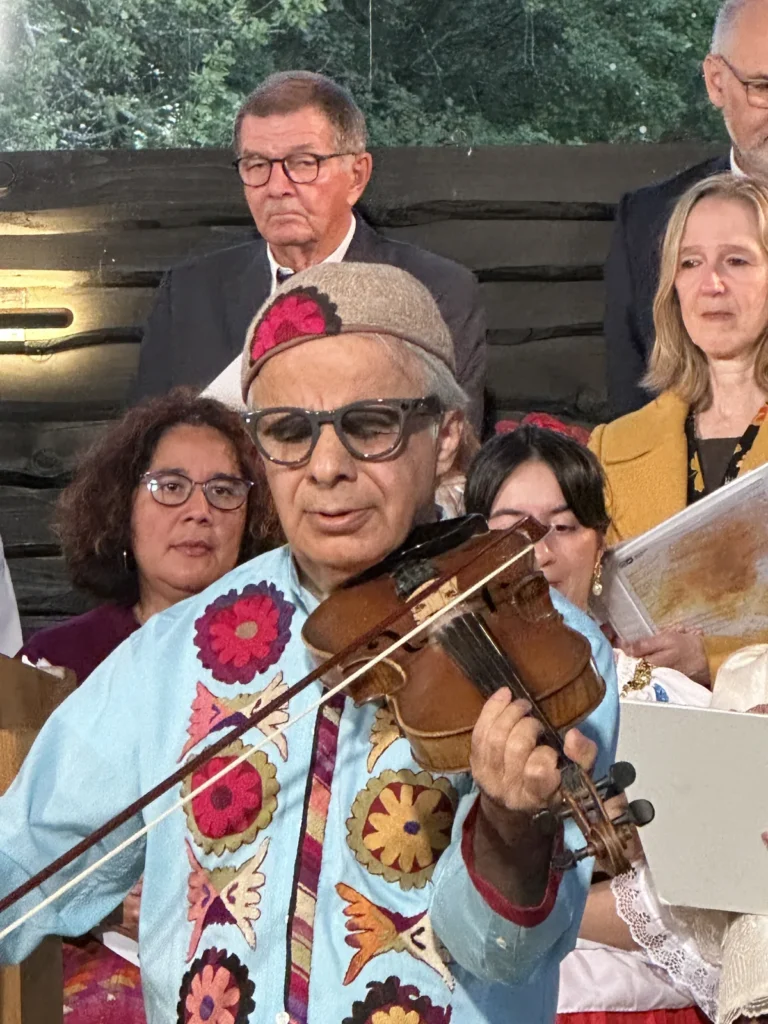 A musician in embroidered traditional costume plays the violin, behind them are the singers of the choir